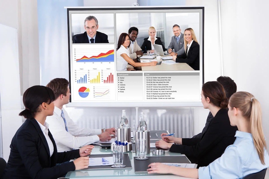 five people sit at conference table looking at a large display with online meeting participants. 